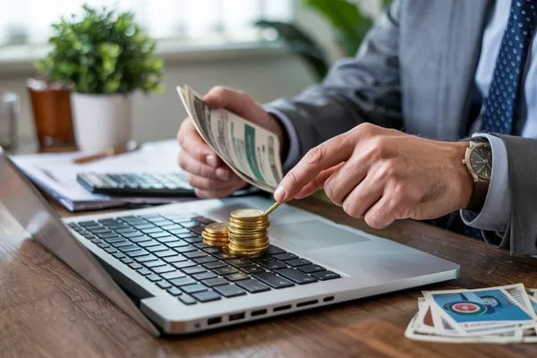 Homme en costume tenant des billets et une pile de pièces dorées posées sur le clavier d’un ordinateur portable.