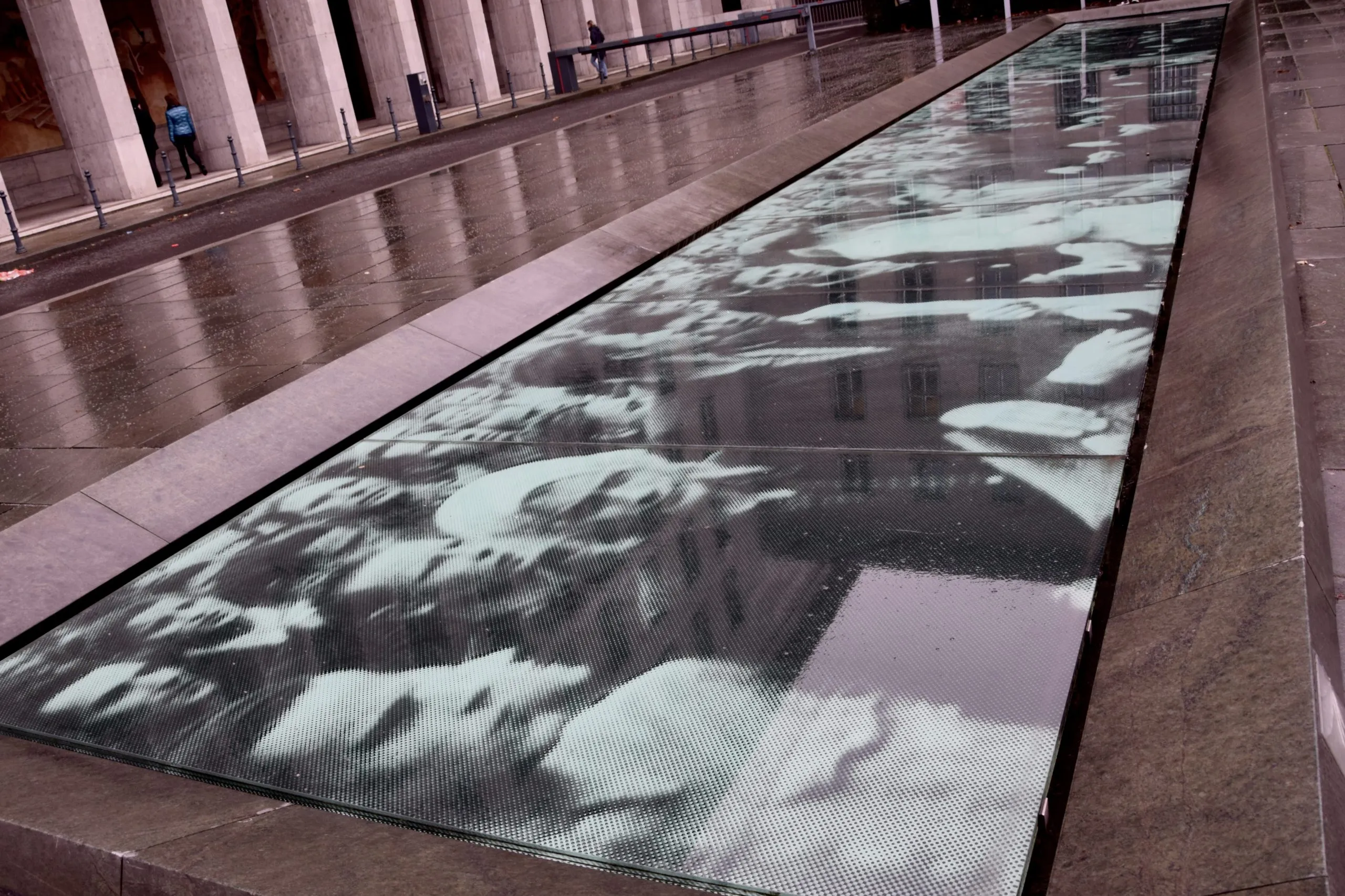 Dalle de verre encastrée dans une place pavée mouillée, reproduisant une photo de foule en noir et blanc, devant une colonnade